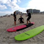 guy shows two people how to properly stand on a surfboard while on land