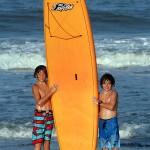 two young boys pose next to orange surfboard on beach