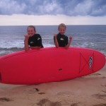boy and girl pose next to surfboard with big smiles and cloudy skies in the back